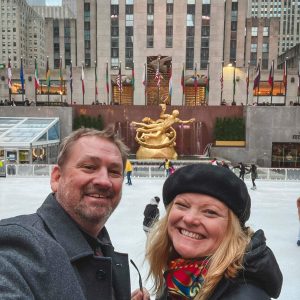 Dave and Deb from The Planet D smiling in front of Rockefeller Center at Christmas in New York City.